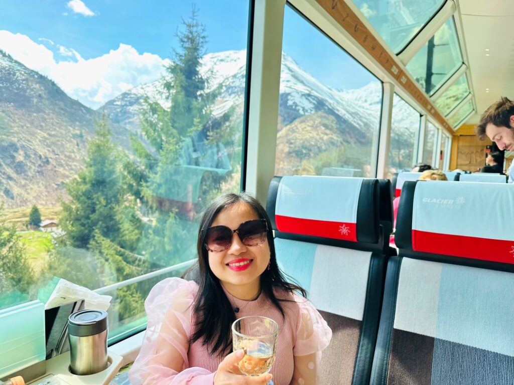 Tôi trên tàu ngắm cảnh Thuỵ Sĩ tay cầm ly vang trắng, thưởng thức cảnh núi tuyết Alps