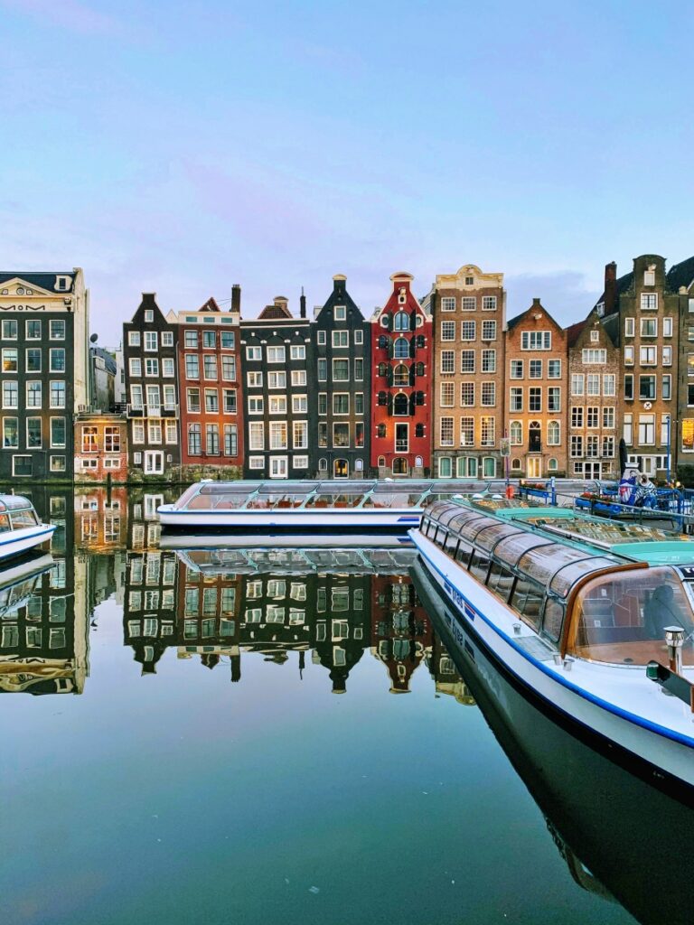 Dãy nhà nghiêng bên kênh Damrak Amsterdam Hà Lan, gần ga trung tâm, phản chiếu xuống mặt nước – điểm check-in nổi tiếng của du khách quốc tế